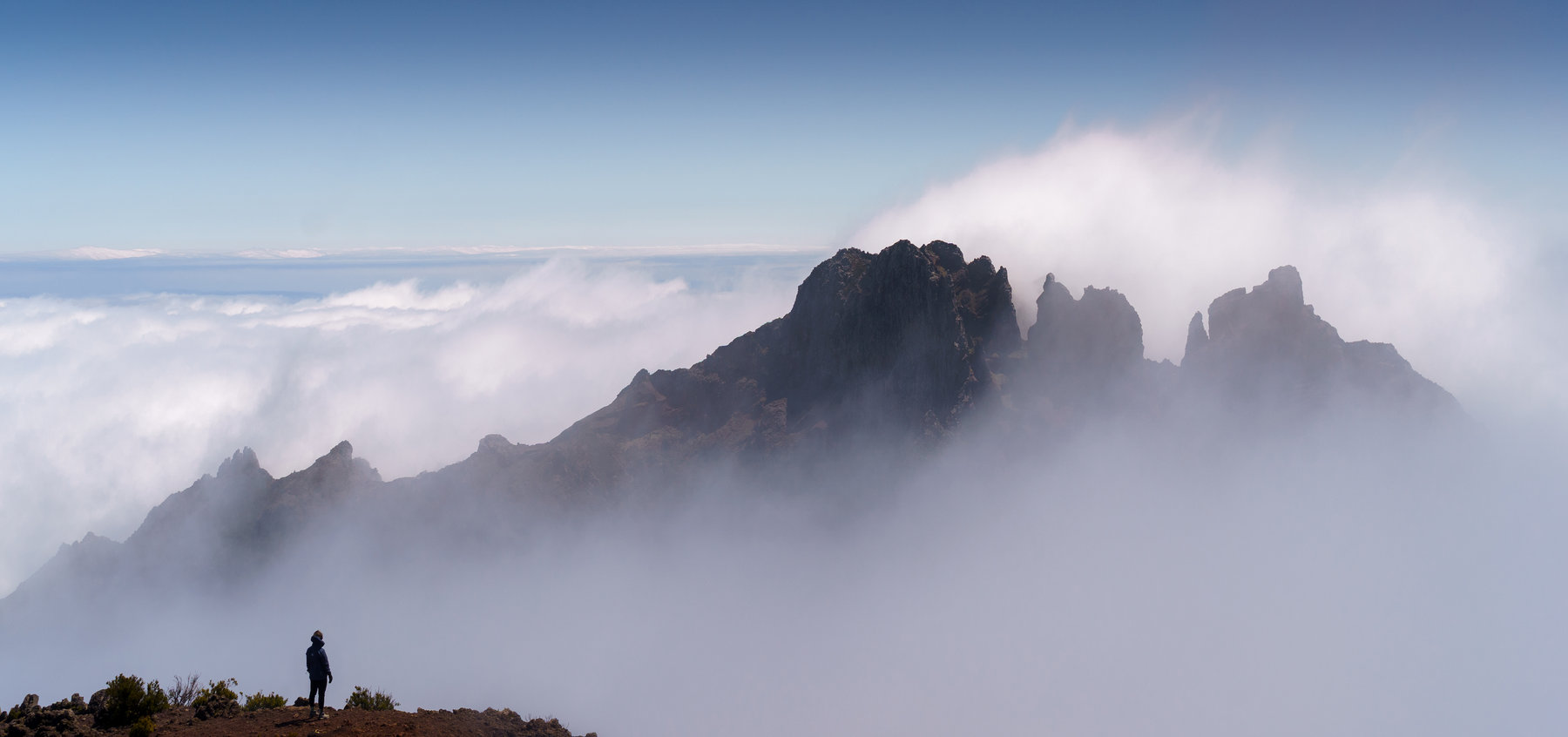 A lone figure on a ridge in front of mountain peaks rising out of the clouds